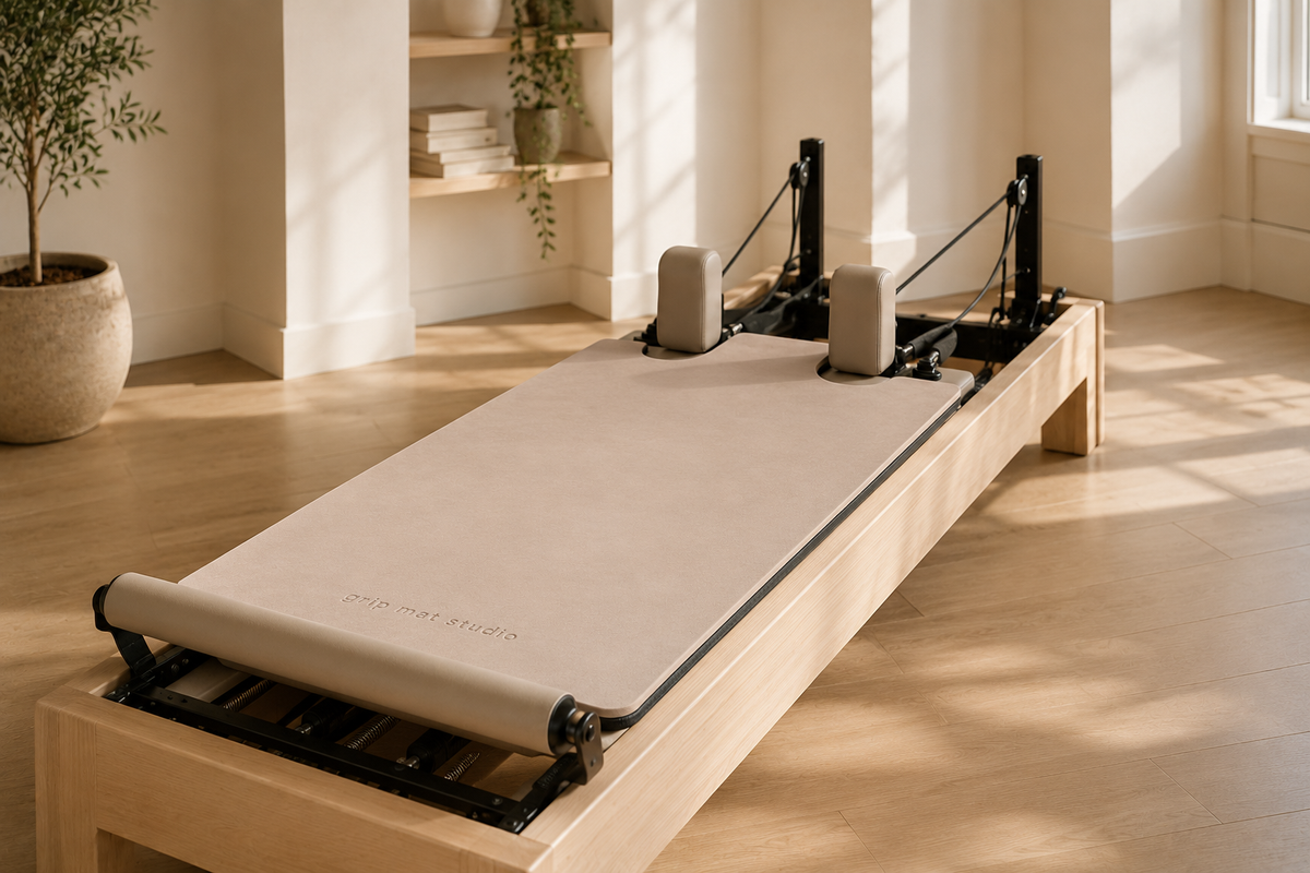 A Pilates reformer machine with a beige fabric slide and black metal frame, positioned on a light wood floor in a bright, modern room with warm sunlight and window shadows.