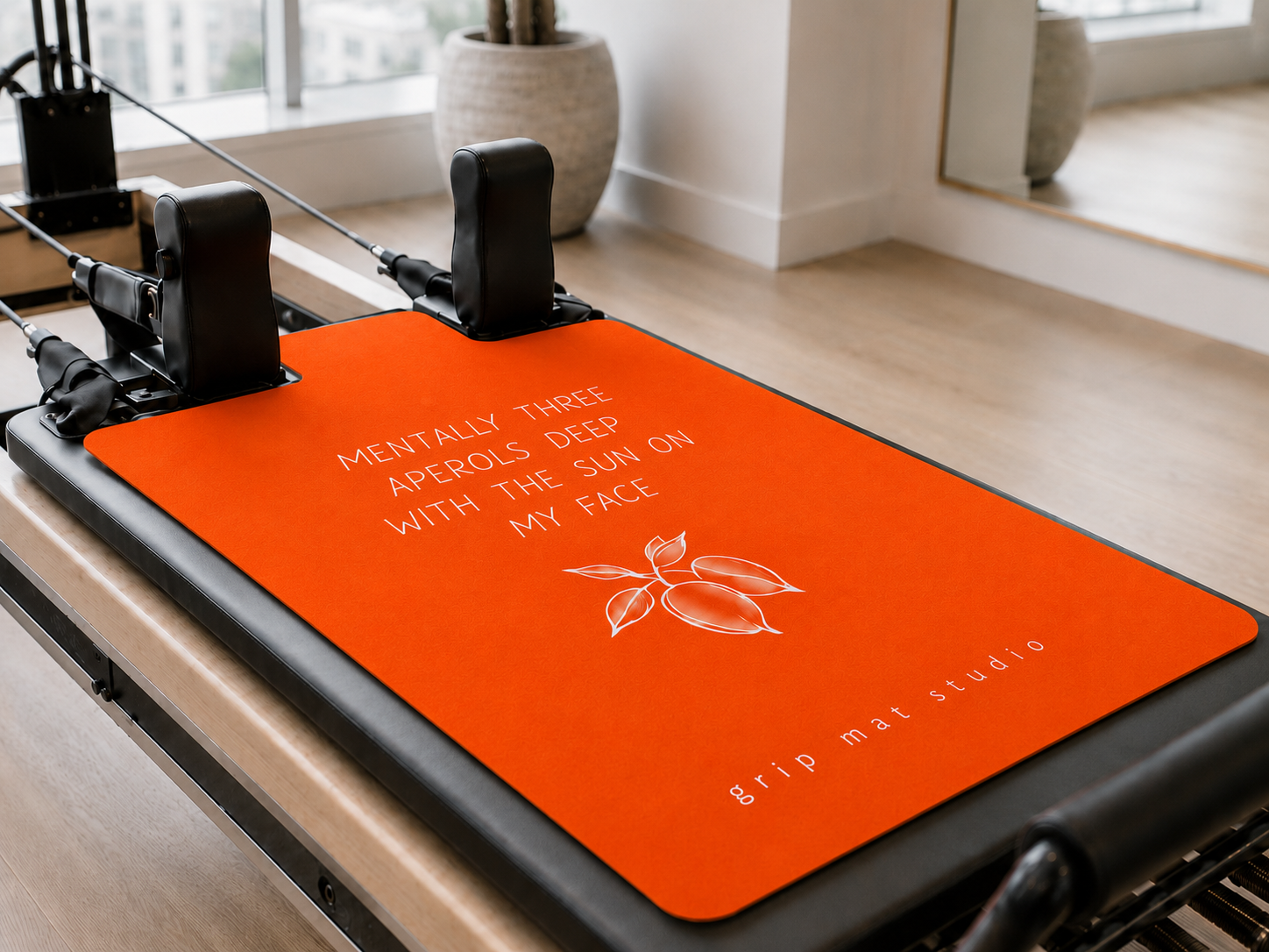 Bright orange Grip Mat Studio Pilates reformer grip mat with “Mentally three Aperols deep with the sun on my face” design in a modern studio.