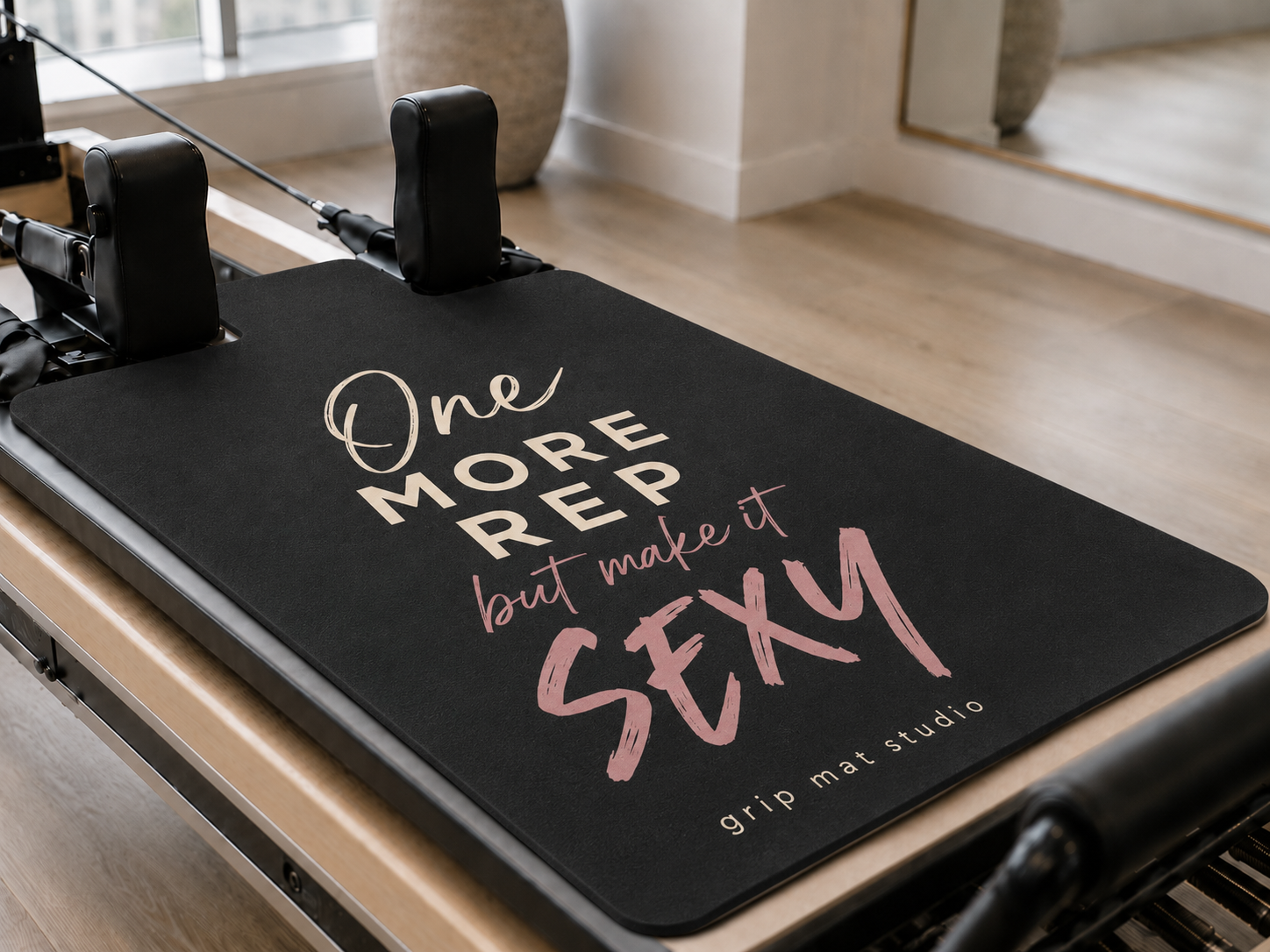 A Pilates reformer machine with a black grip mat featuring the motivational quote 'One MORE REP but make it SEXY' in white and pink lettering, with 'grip mat studio' printed at the bottom.