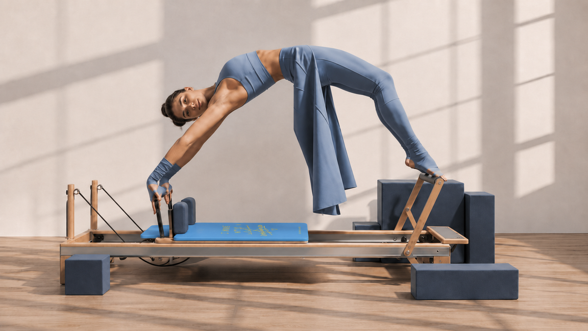 A woman in a blue workout set performs a yoga pose on a Pilates reformer machine in a bright, modern studio with warm sunlight and window shadows.