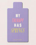 A lavender grip mat with the text 'MY THERAPY HAS SPRINGS' in white, pink, and yellow lettering, displayed on a light beige background.