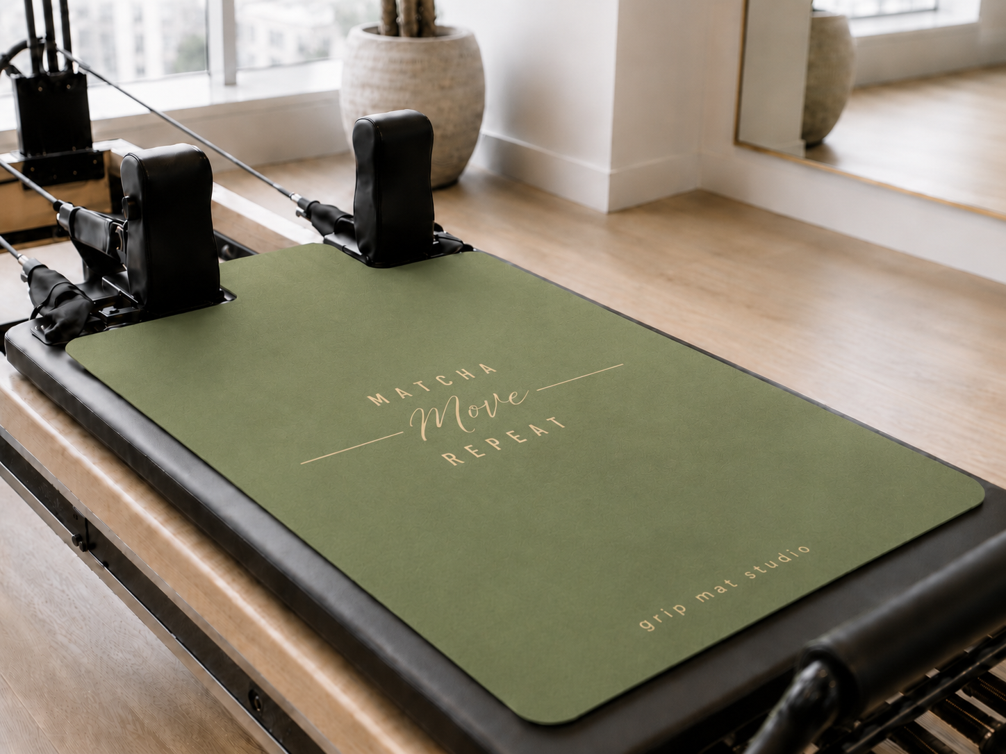 An olive green Pilates reformer mat with gold foil lettering reading 'MATCHA Move REPEAT' and 'grip mat studio', shown on a wooden reformer machine in a bright, modern studio.