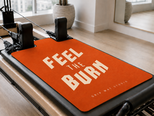 An orange Pilates reformer grip mat with the words 'FEEL THE BURN' in white, displayed on a reformer machine in a bright, modern studio.