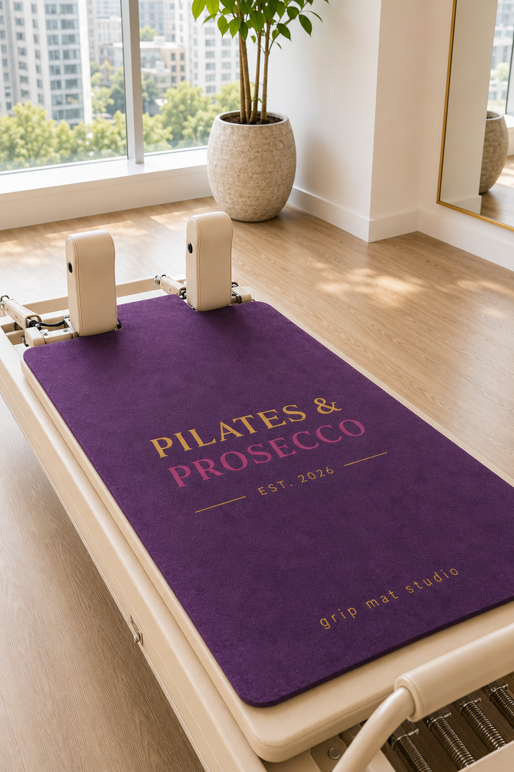A purple Pilates reformer mat with custom printed text reading 'PILATES & PROSECCO EST. 2026' and 'grip mat studio' in gold and pink, shown on a cream-colored reformer machine in a bright, modern studio with a window and potted plant.