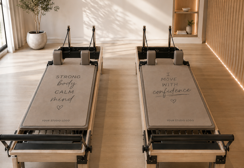Two Pilates reformer machines placed side by side in a bright, modern studio with light wood floors and warm sunlight streaming in. Each reformer has a beige mat with black and gray components, and printed motivational text is visible on the mats.