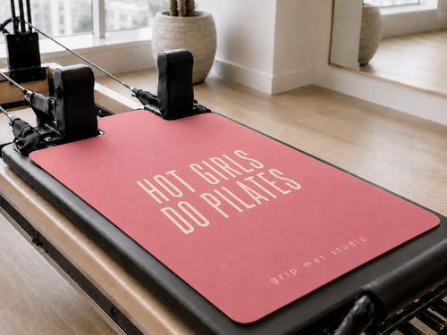 A pink Pilates reformer mat with the text 'HOT GIRLS DO PILATES' in cream lettering, placed on a black and wood Pilates reformer machine in a bright, modern studio.