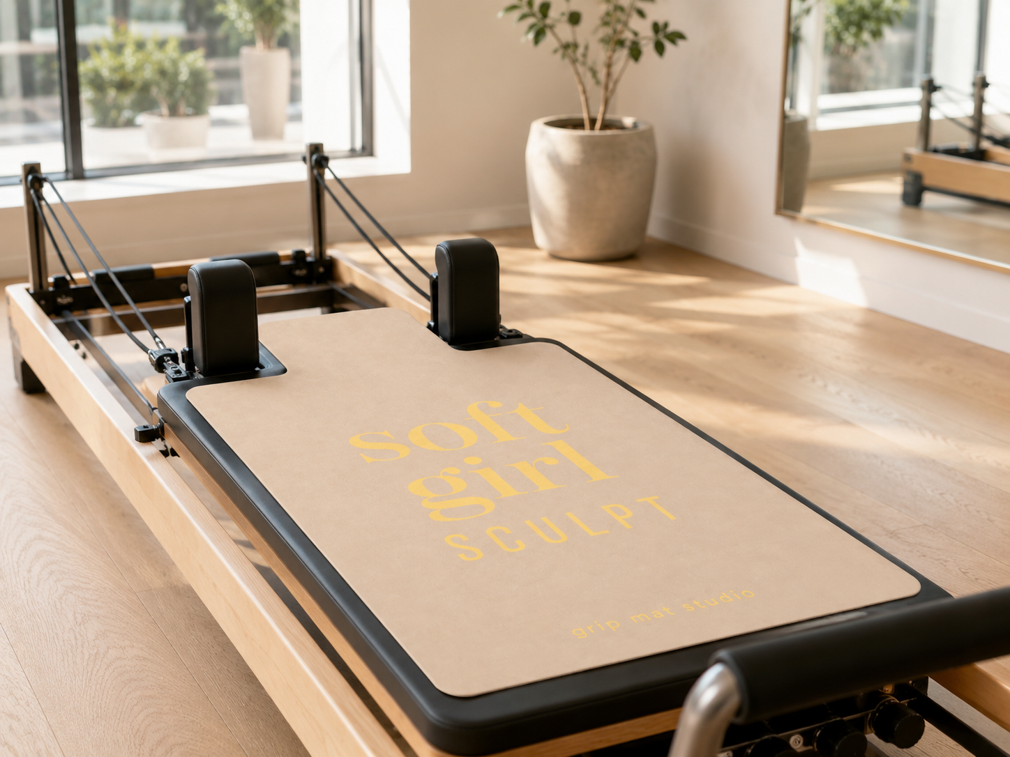A Pilates reformer machine in a bright studio with wood floors and windows. The pad on the reformer reads 'soft girl SCULPT' in yellow text.