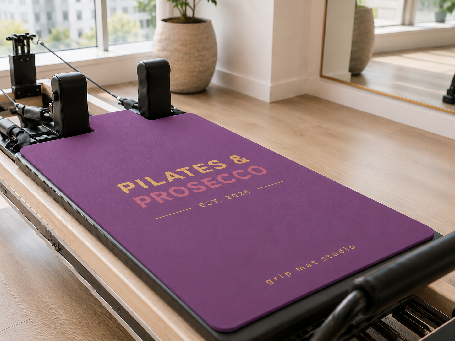 A purple Pilates reformer mat with 'PILATES & PROSECCO EST. 2026' printed in pink and gold, shown on a wooden reformer machine in a bright, modern studio.