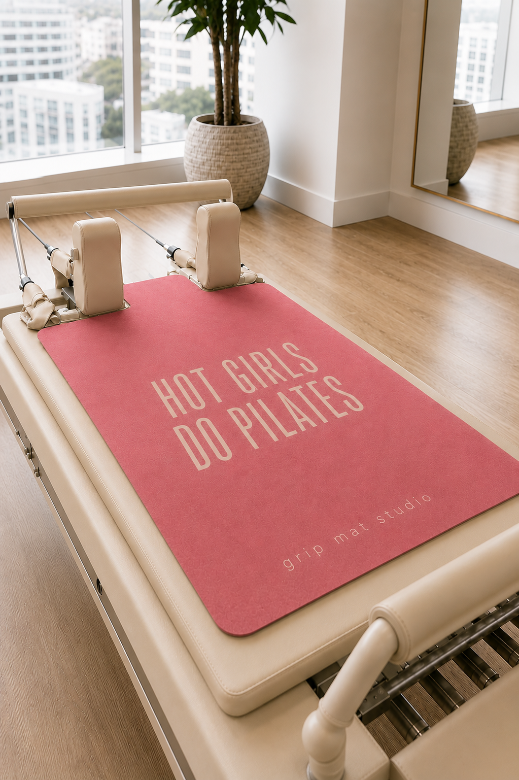 Pink "Hot Girls Do Pilates" reformer grip mat displayed on a beige Pilates reformer in a bright, modern studio with floor-to-ceiling windows, light wood floors, and neutral décor. The premium suede mat features a minimalist cream typographic design and Grip Mat Studio branding, creating a clean, luxury Pilates aesthetic.