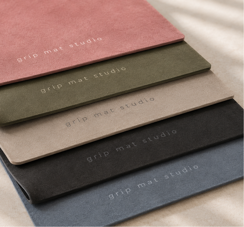 Four Grip Mat Studio textured desk mats stacked diagonally on a light fabric background, with text callouts pointing to each color variant: dusty pink, olive green, taupe, and charcoal gray.