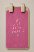 A pink grip mat with a beige border and two beige handle grips at the top, featuring the white text 'LOVE FLOW PILATES' and small heart illustrations on the front. The brand 'grip mat studio' is printed at the bottom.
