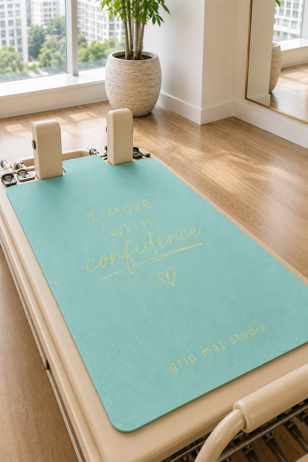 A turquoise Pilates reformer mat with gold lettering reading 'I MOVE WITH confidence' and 'grip mat studio', placed on a beige reformer machine in a bright room with wood floors and a window.