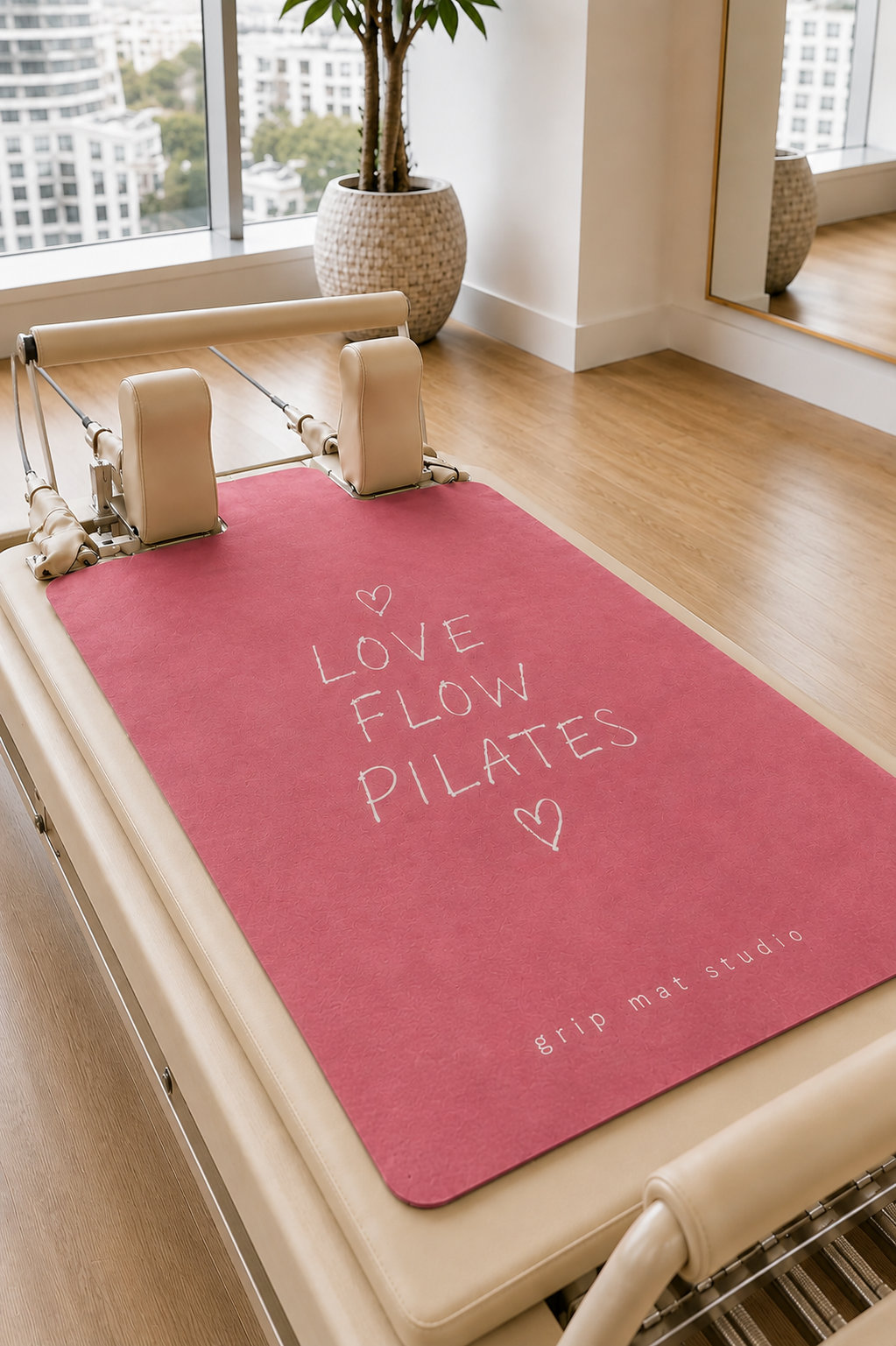 Pink "Hot Girls Do Pilates" reformer grip mat displayed on a beige Pilates reformer in a bright, modern studio with floor-to-ceiling windows, light wood floors, and neutral décor. The premium suede mat features a minimalist cream typographic design and Grip Mat Studio branding, creating a clean, luxury Pilates aesthetic.