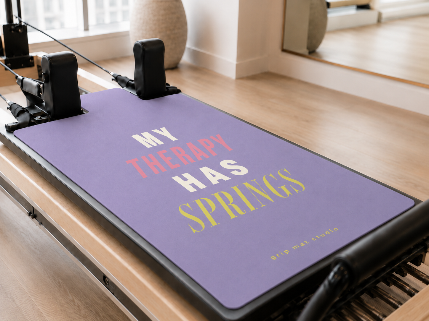 A Pilates reformer machine in a bright studio setting, featuring a lavender purple mat printed with the quote 'MY THERAPY HAS SPRINGS' in white, pink, and yellow text.