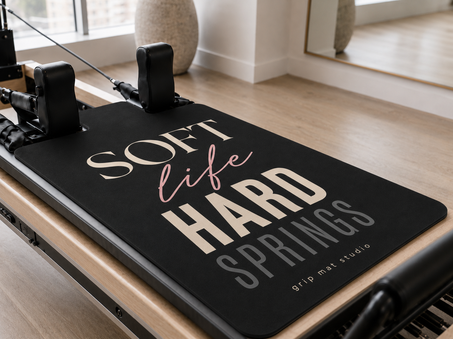 A Pilates reformer machine with a black grip mat featuring the text 'SOFT life HARD SPRINGS' and 'grip mat studio' in cream and pink lettering, set in a bright, modern studio with light wood floors.