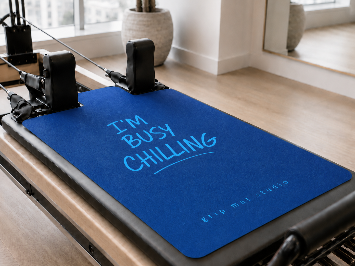 A blue grip mat on a reformer machine with the text 'I'M BUSY CHILLING' in light blue, photographed in a bright indoor studio setting.