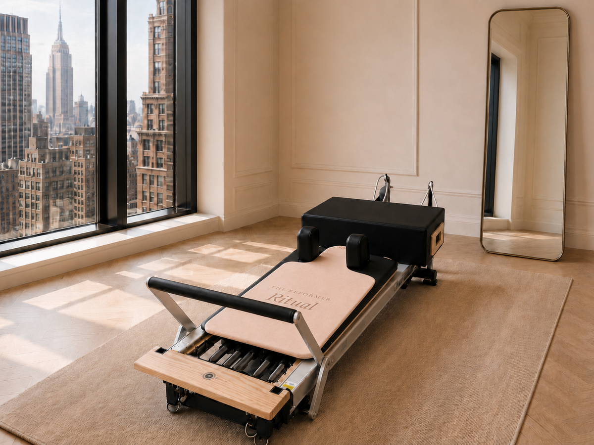 A Pilates reformer machine with a black and natural wood frame and a beige fabric slide, set on a beige rug in a bright room with a large window showing a city skyline. A full-length mirror is visible on the right.