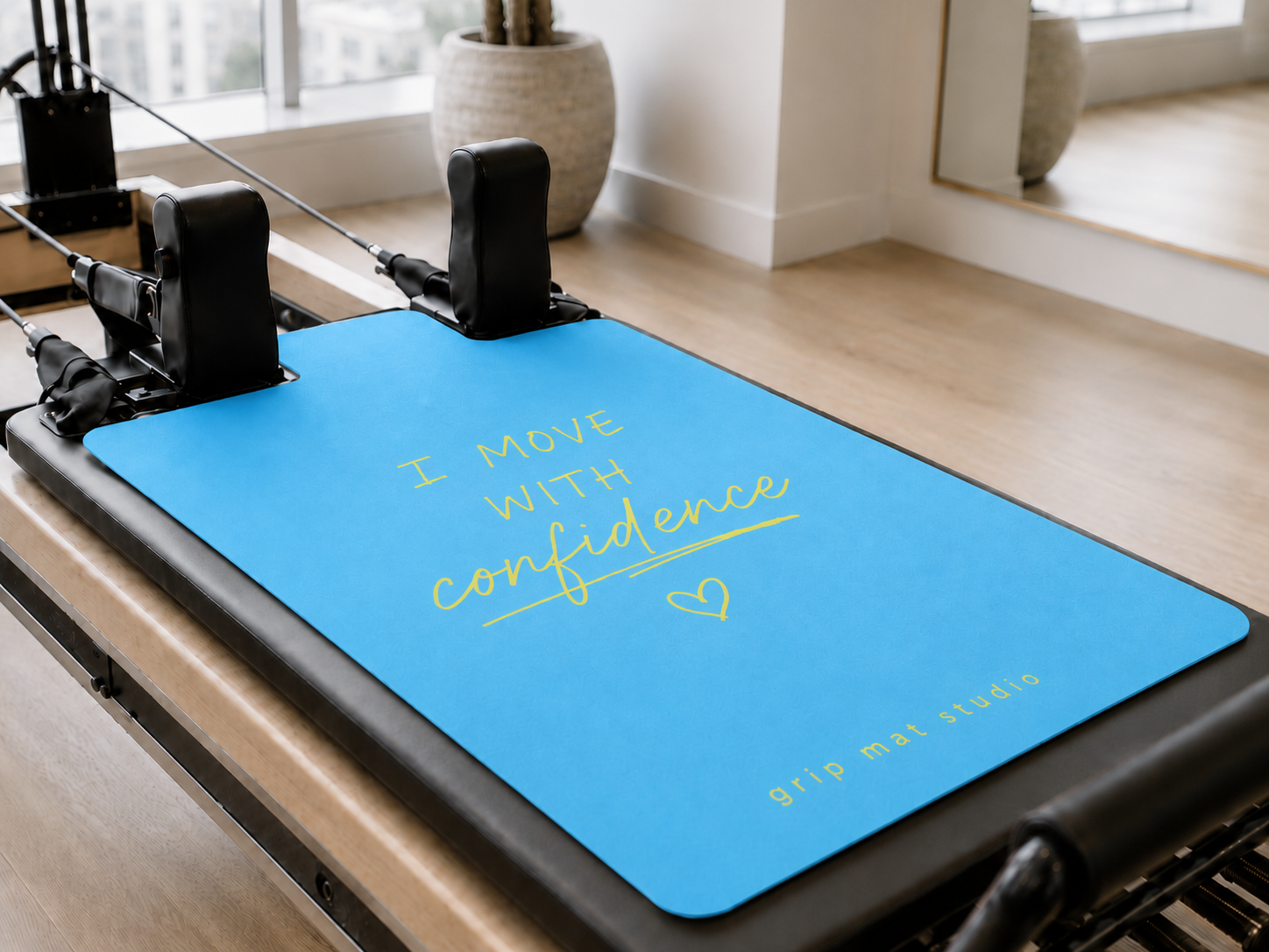 Light blue “I Move With Confidence” Pilates reformer grip mat with yellow lettering on a modern studio reformer.