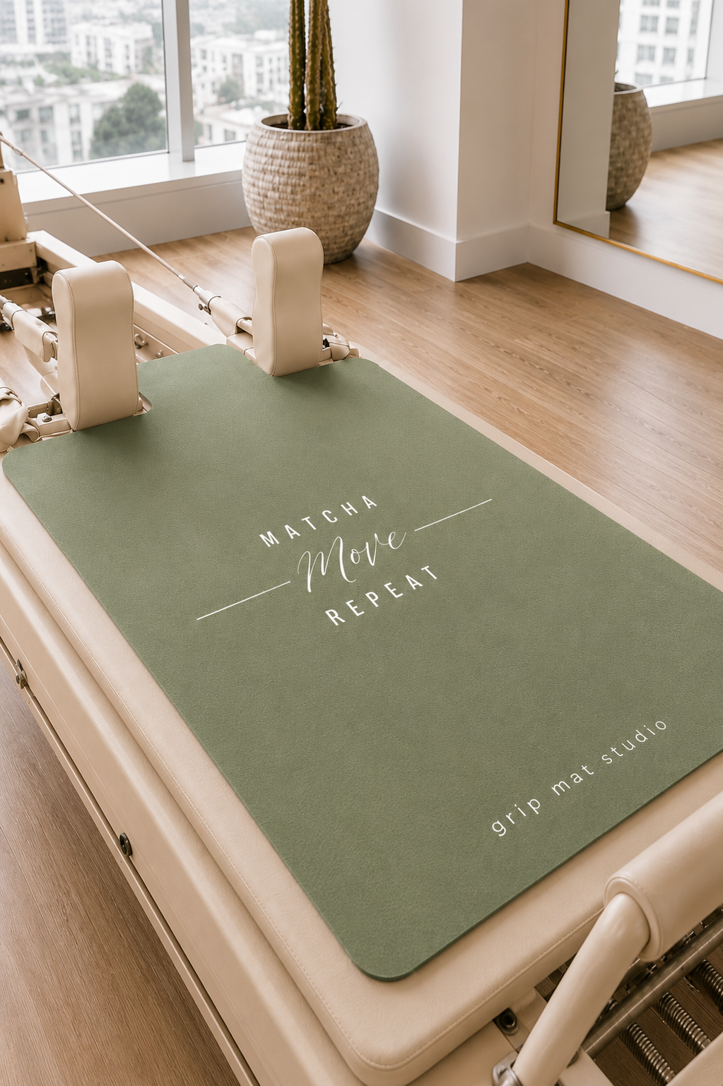 An olive green Pilates reformer mat with white text reading 'MATCHA Move REPEAT' and 'grip mat studio' in the corner, placed on a cream-colored reformer machine in a bright, modern studio with wood floors and a window.