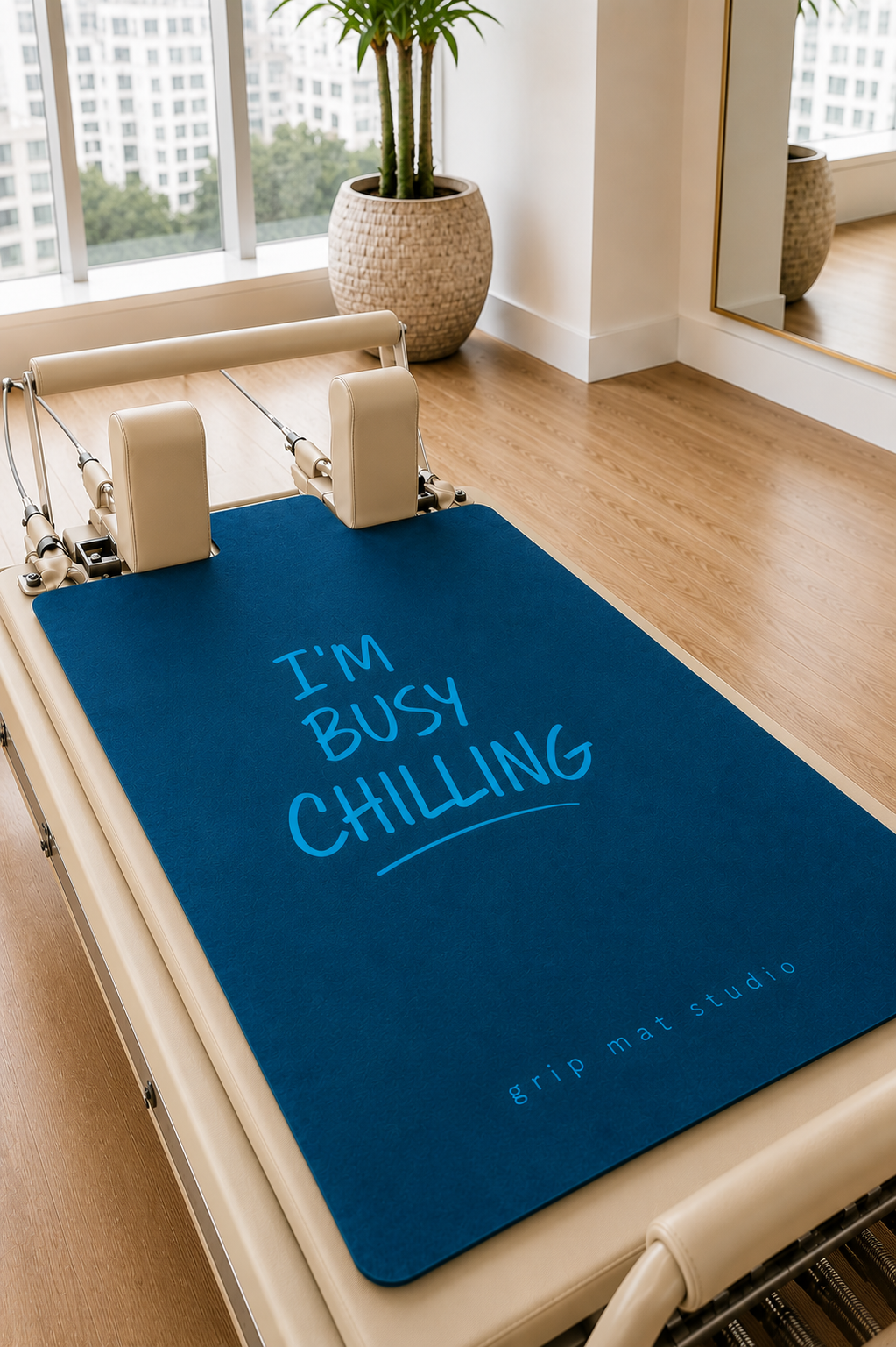 A blue Pilates reformer mat with the text 'I'M BUSY CHILLING' in light blue, displayed on a cream-colored reformer machine in a bright, modern studio with wood floors and a potted plant.