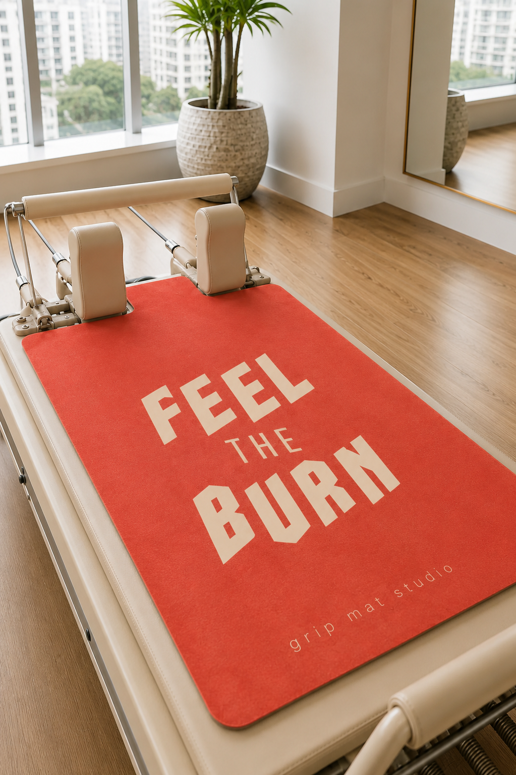 A red Pilates reformer mat with the words 'FEEL THE BURN' in white text, placed on a cream-colored reformer machine in a bright studio with wood floors and a window.