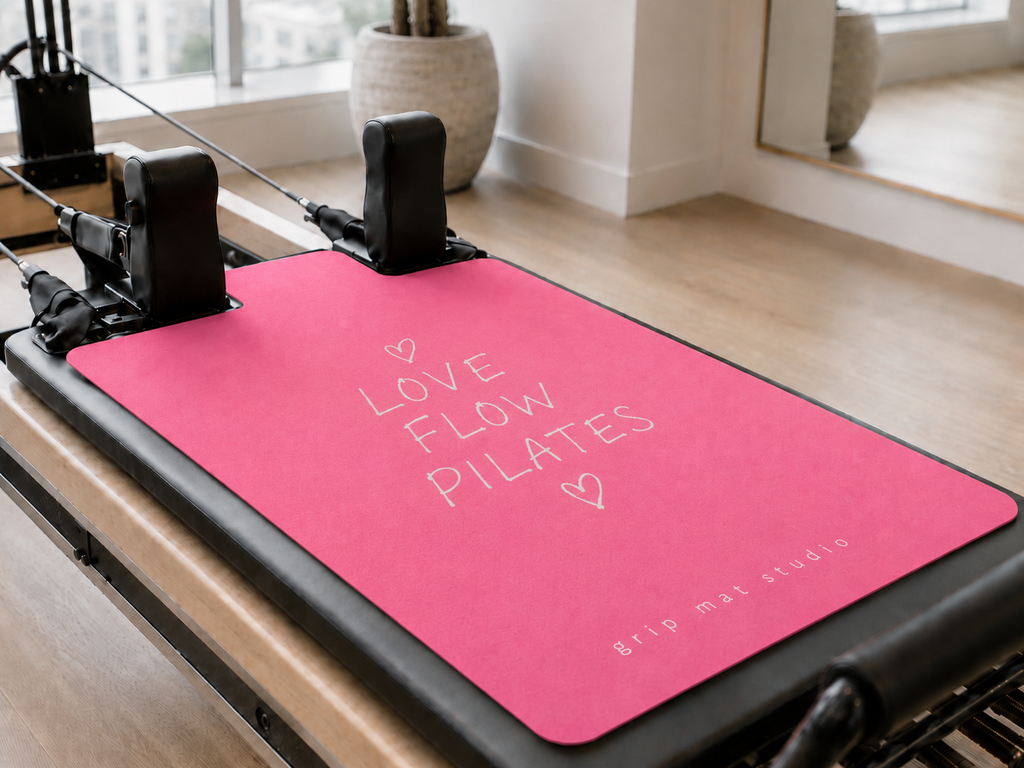 A pink Pilates reformer mat with white text reading 'LOVE FLOW PILATES' and 'grip mat studio' in the corner, placed on a Pilates reformer machine in a bright, modern studio.